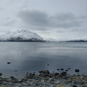 Snow-capped mountains reflected in calm water, with a rocky shoreline in the foreground under a cloudy sky.