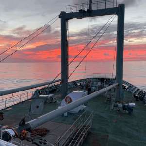 Bow of a ship at sea during sunset, with a pole and machinery visible, under a sky filled with red and orange clouds.