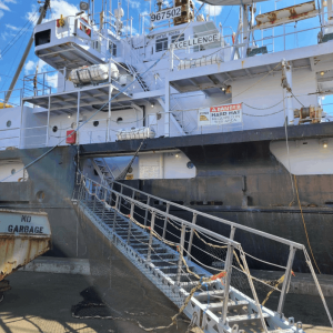 Large docked ship named "Excellence" with visible stairway and signs. Blue sky and ropes in the background.